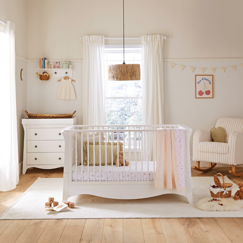 Light and airy neutral baby nursery featuring a white wooden crib with cherry print bedding, a matching white changing dresser, soft rocking chair, natural woven accents, and warm wooden flooring, styled in a calm Scandinavian-inspired nursery design.